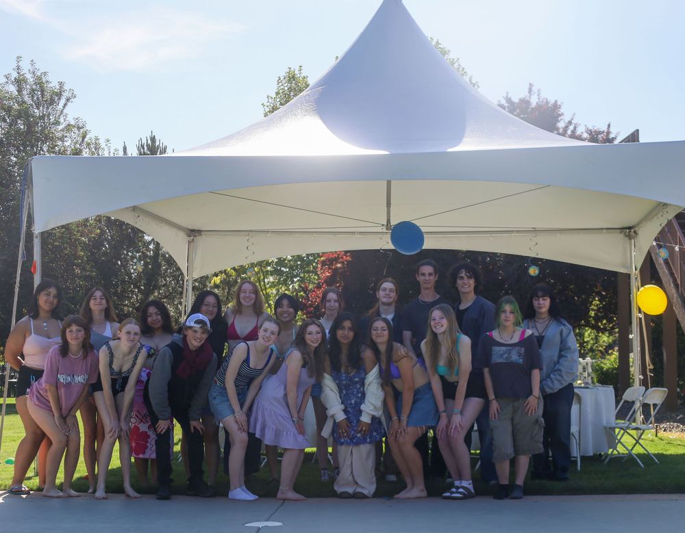 A group gathers under a white tent canopy with yellow and blue balloons during an outdoor event.