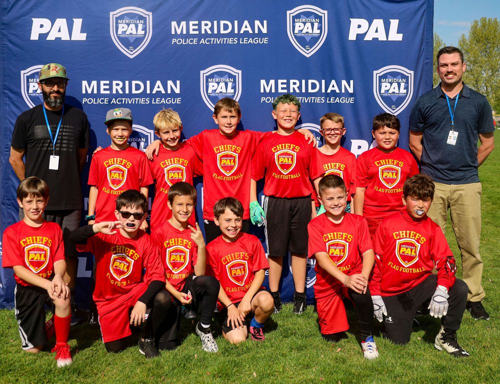 Youth soccer team in red jerseys poses for group photo with coaches in front of blue Meridian PAL banner on sunny field.