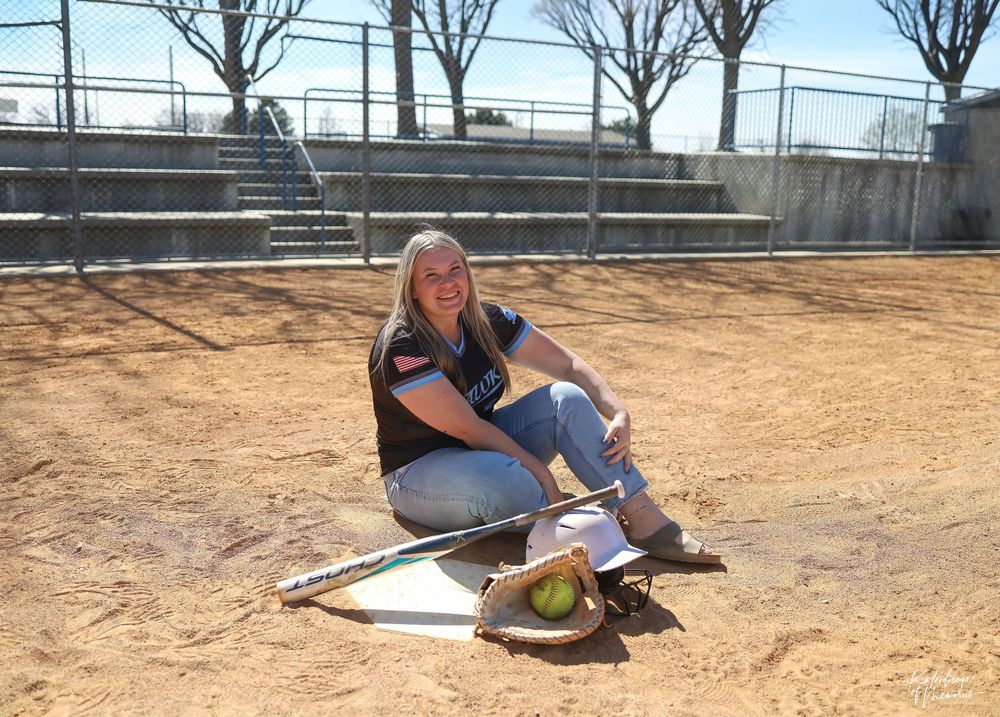 Young female softball player sitting on home plate with bat and glove on a dirt baseball field outdoors.