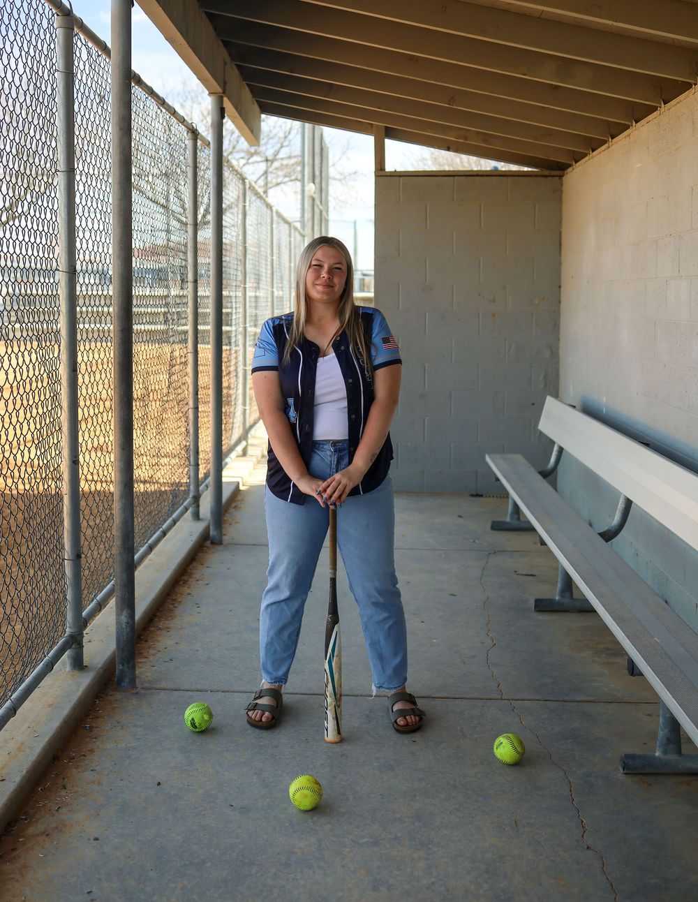Young female softball player stands in dugout holding bat, with tennis balls at her feet, wearing team jersey and jeans.