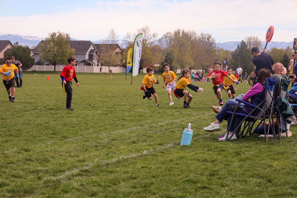 Children playing flag football on a grass field while spectators watch from sideline chairs on an autumn afternoon.