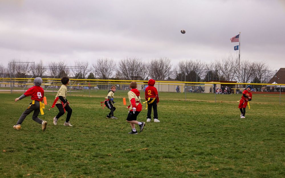 A group plays flag football on a grassy field on an overcast day with American flag in background.