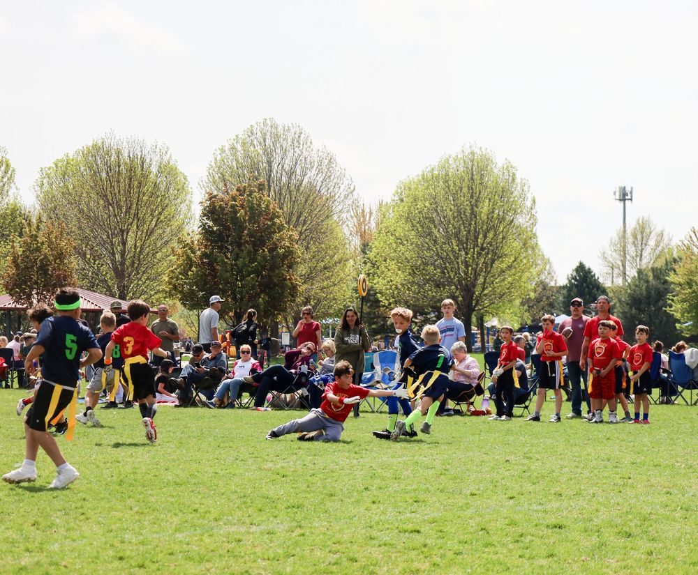 Outdoor rugby match with players running and tackling on a green field while spectators watch from the sidelines.