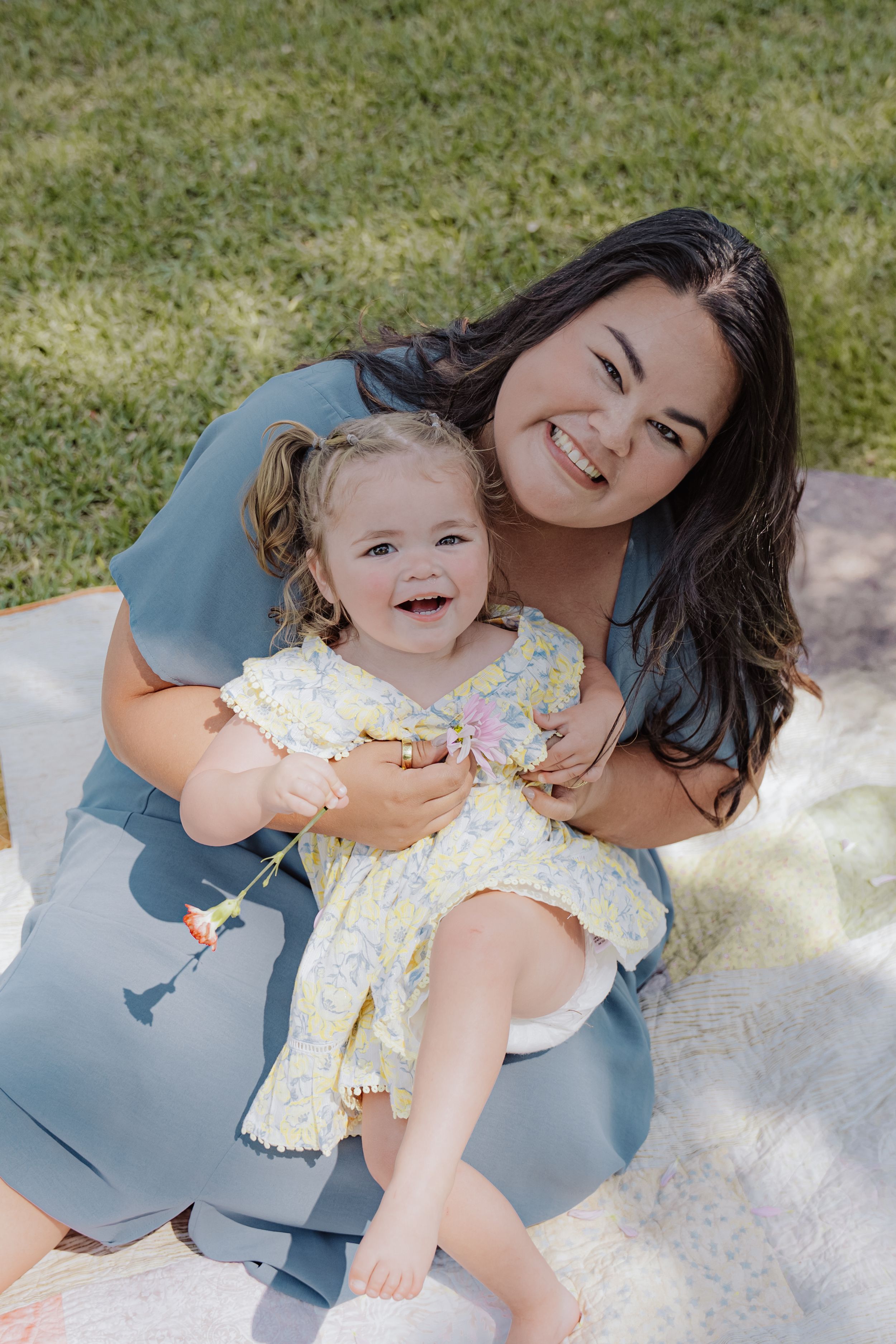 Mom and daughter sit together on a blanket outdoors, sharing a joyful moment in casual attire on a sunny day.