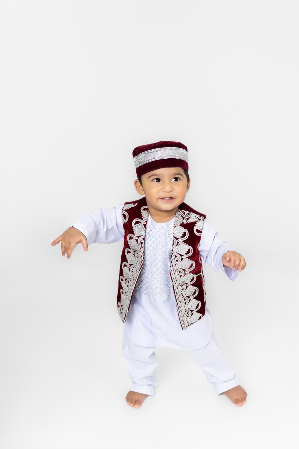 Baby wearing traditional white and burgundy Middle Eastern cultural attire with embroidered vest and matching cap.