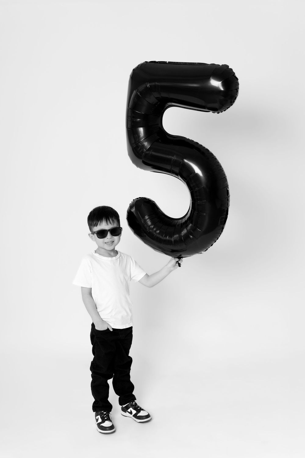 A young child in sunglasses and casual attire poses with a large black number 5 balloon against a white background.