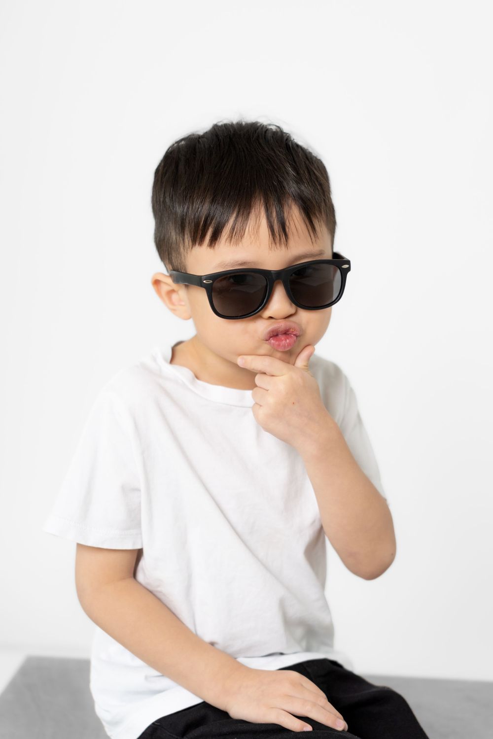 A small child in sunglasses and a white t-shirt strikes a thoughtful pose against a white background.