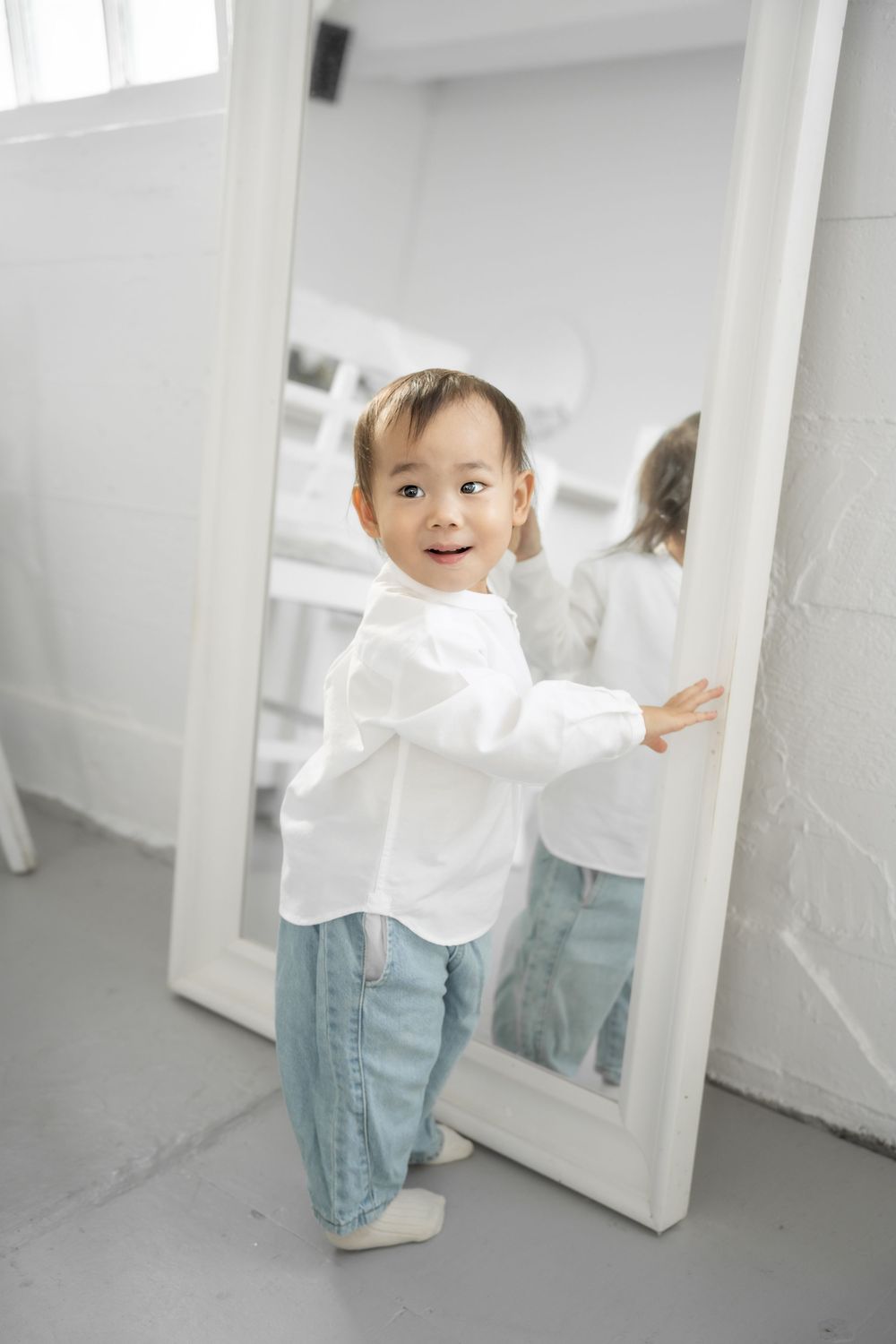 A small child in a white shirt and jeans plays in a bright white room with open doors.