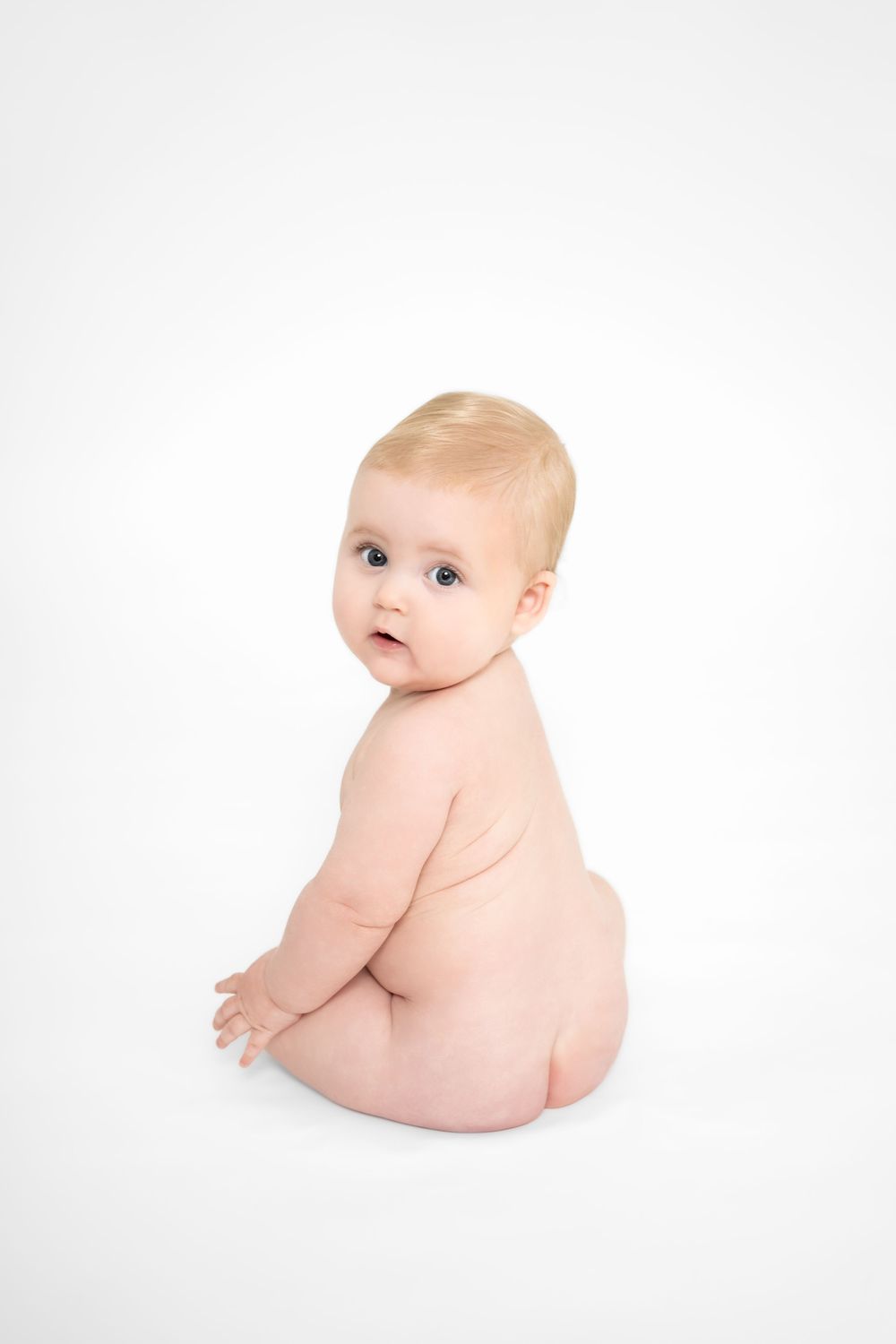 A baby sitting on a white background showing their bare back and looking over their shoulder.