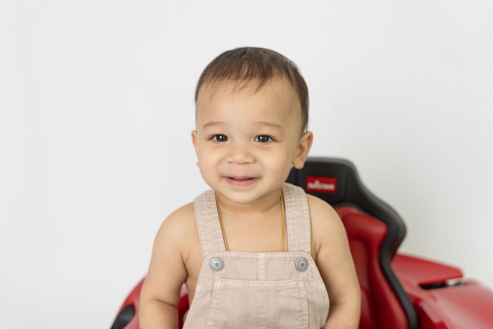 A toddler wearing beige overalls sits in a red chair against a white background.