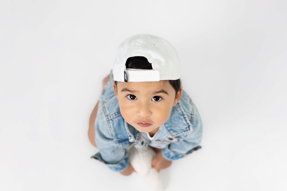 A child wearing a white baseball cap and denim outfit looks up at the camera from a high angle against a white background.