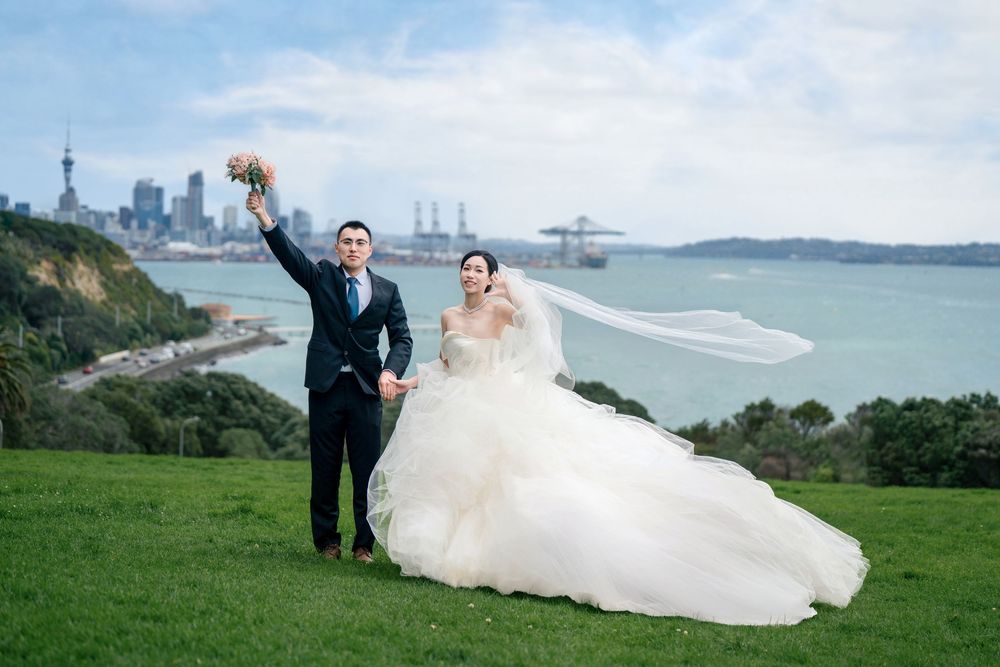 Outdoor pre-wedding photography featuring a happy couple celebrating on a green hill with the Auckland skyline and harbour in the background