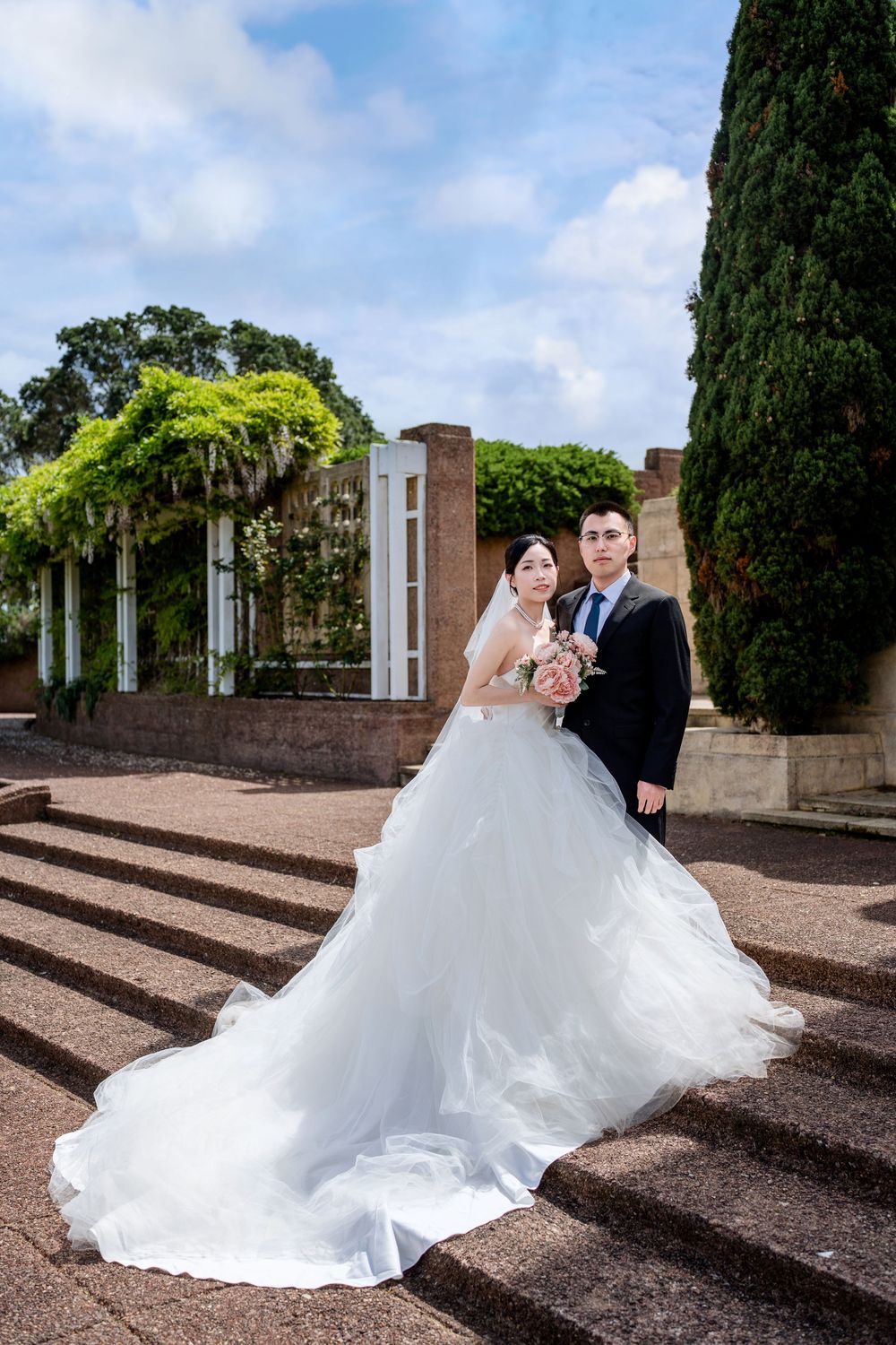 Wide architectural portrait of a bride and groom standing in an elegant brick-paved formal garden.