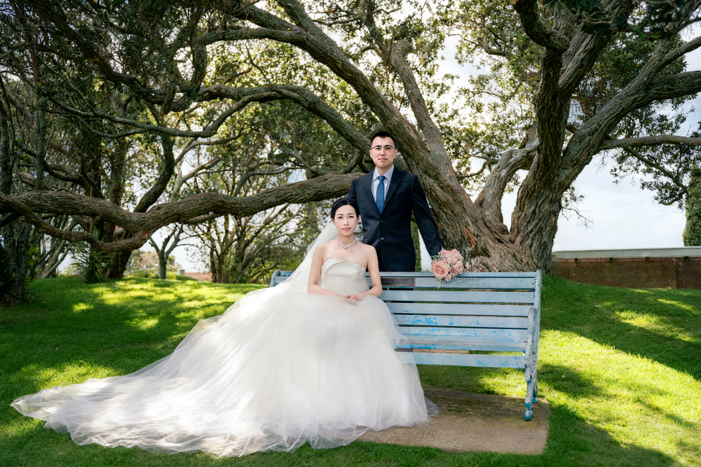 Bride and groom sitting on a classic park bench under large trees for natural Auckland wedding photography.