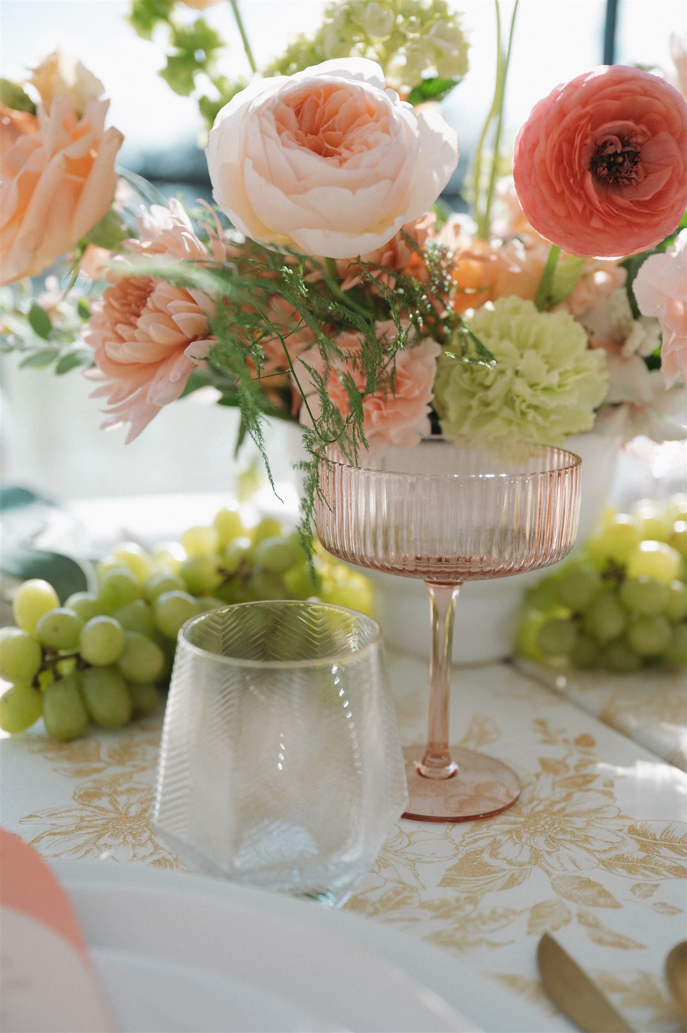 A pink and peach floral arrangement in vintage glassware sits beside fresh green grapes on a delicate tablecloth.