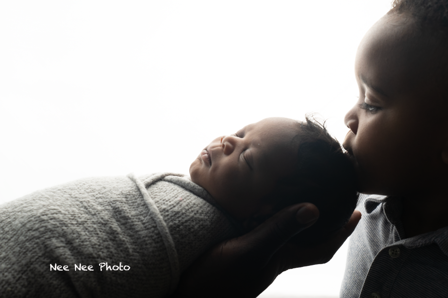 Older sibling gently kissing a swaddled newborn baby in a tender, backlit black-and-white newborn photography session. Embracing Life’s Beau