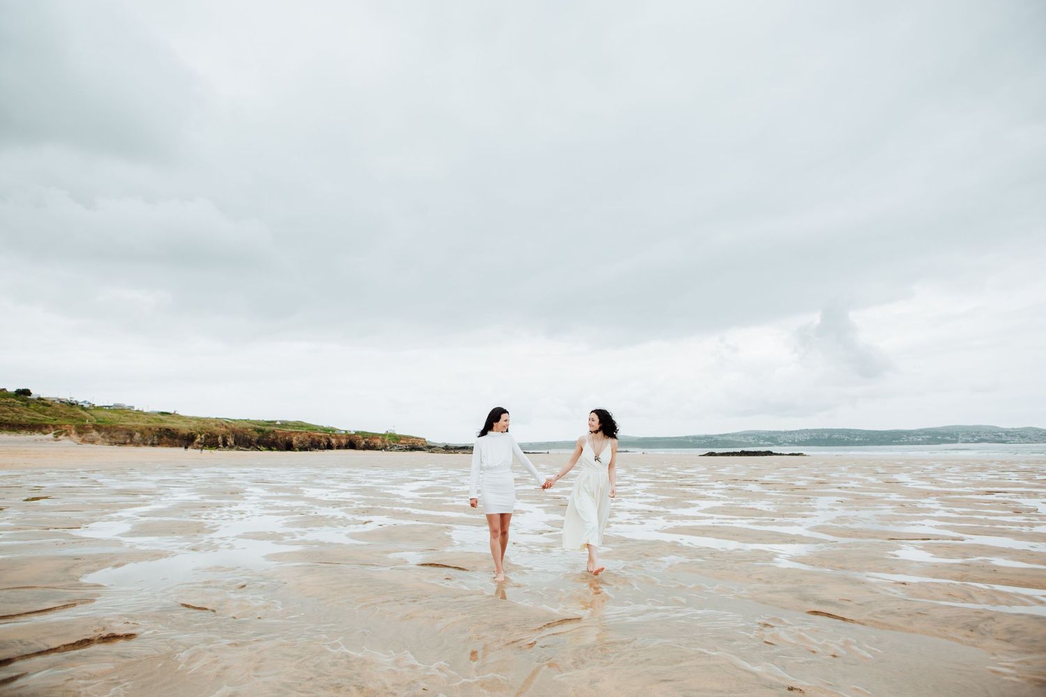 Two figures in white dresses dance joyfully along the seashore.