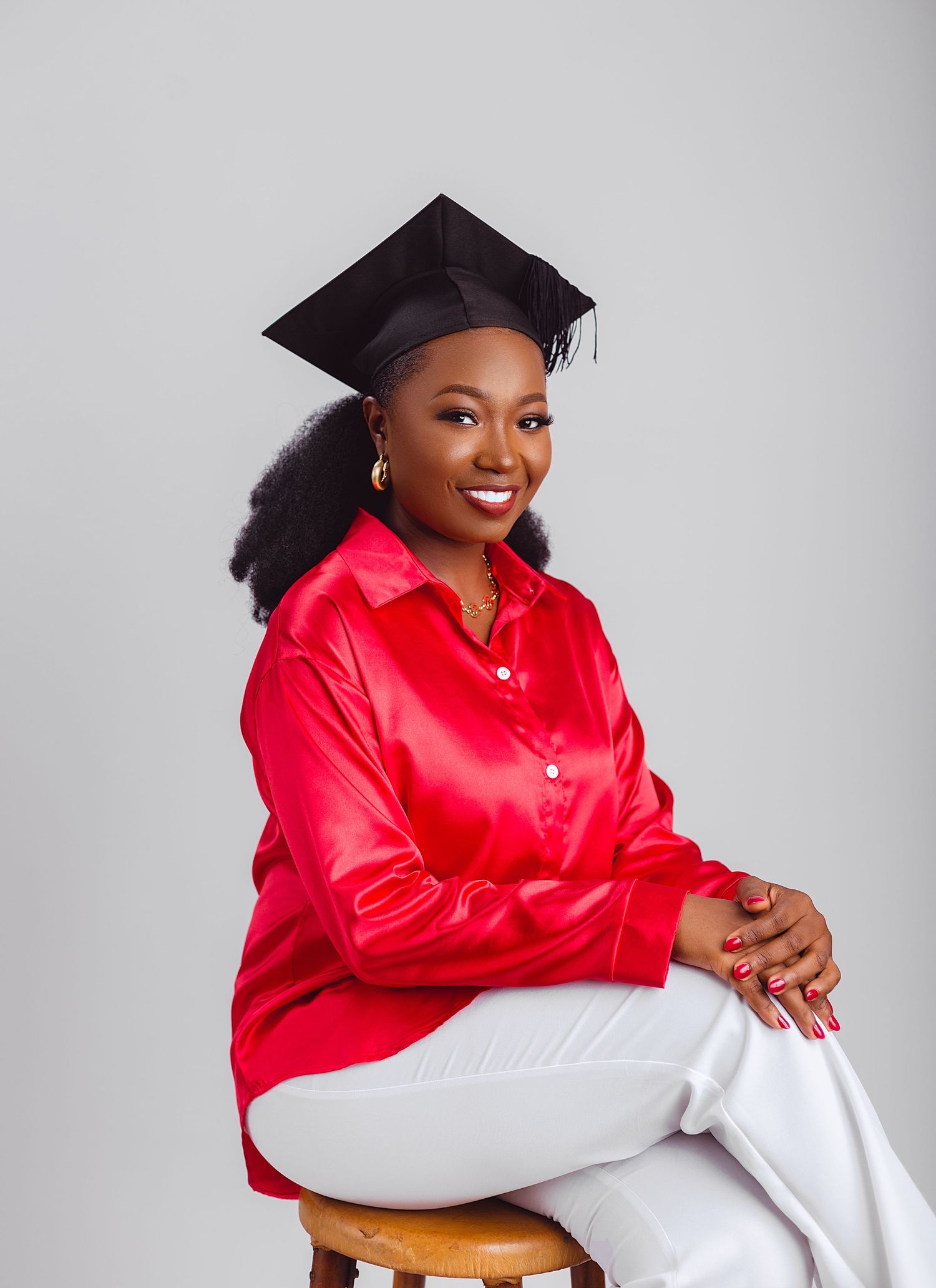 A graduate in a bright red satin shirt and white pants poses with cap and gown against a grey backdrop.