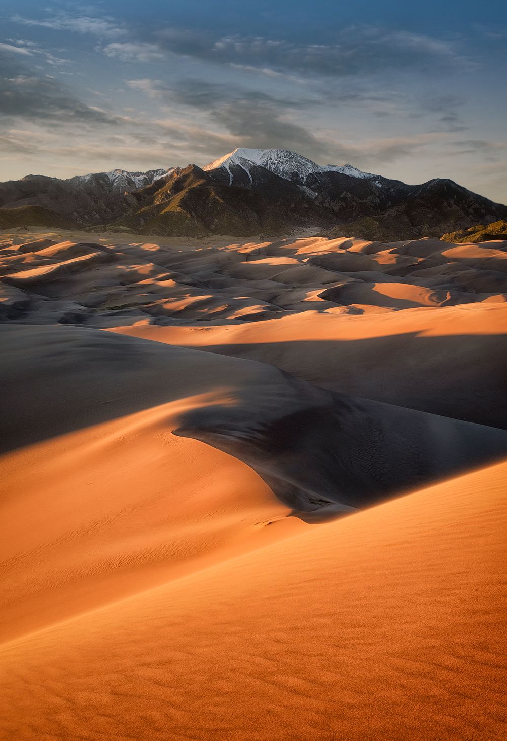 Orange sand dunes contrast with snow-capped mountains under moody sunset skies.