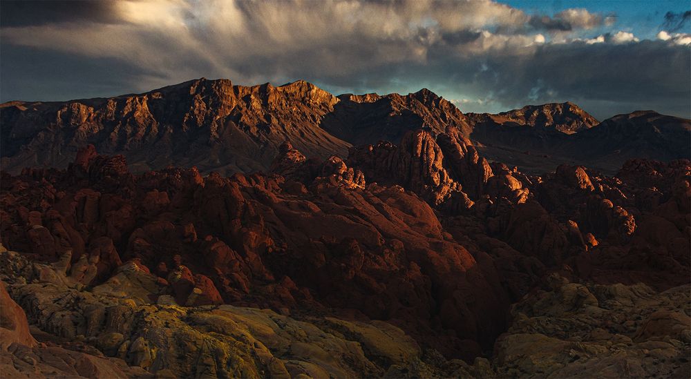 Dramatic mountain landscape at sunset with rugged red rock formations under stormy skies with golden light rays.