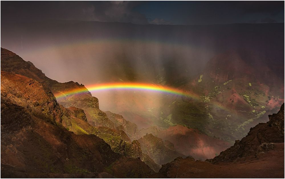 Vibrant rainbow arcs across mountainous canyon landscape during dramatic sunset.