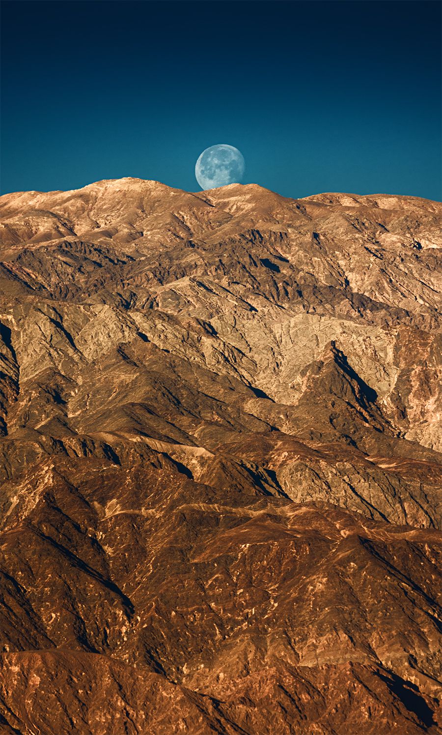 Rugged mountain range under a blue sky with a rising moon casting warm light on rocky terrain.