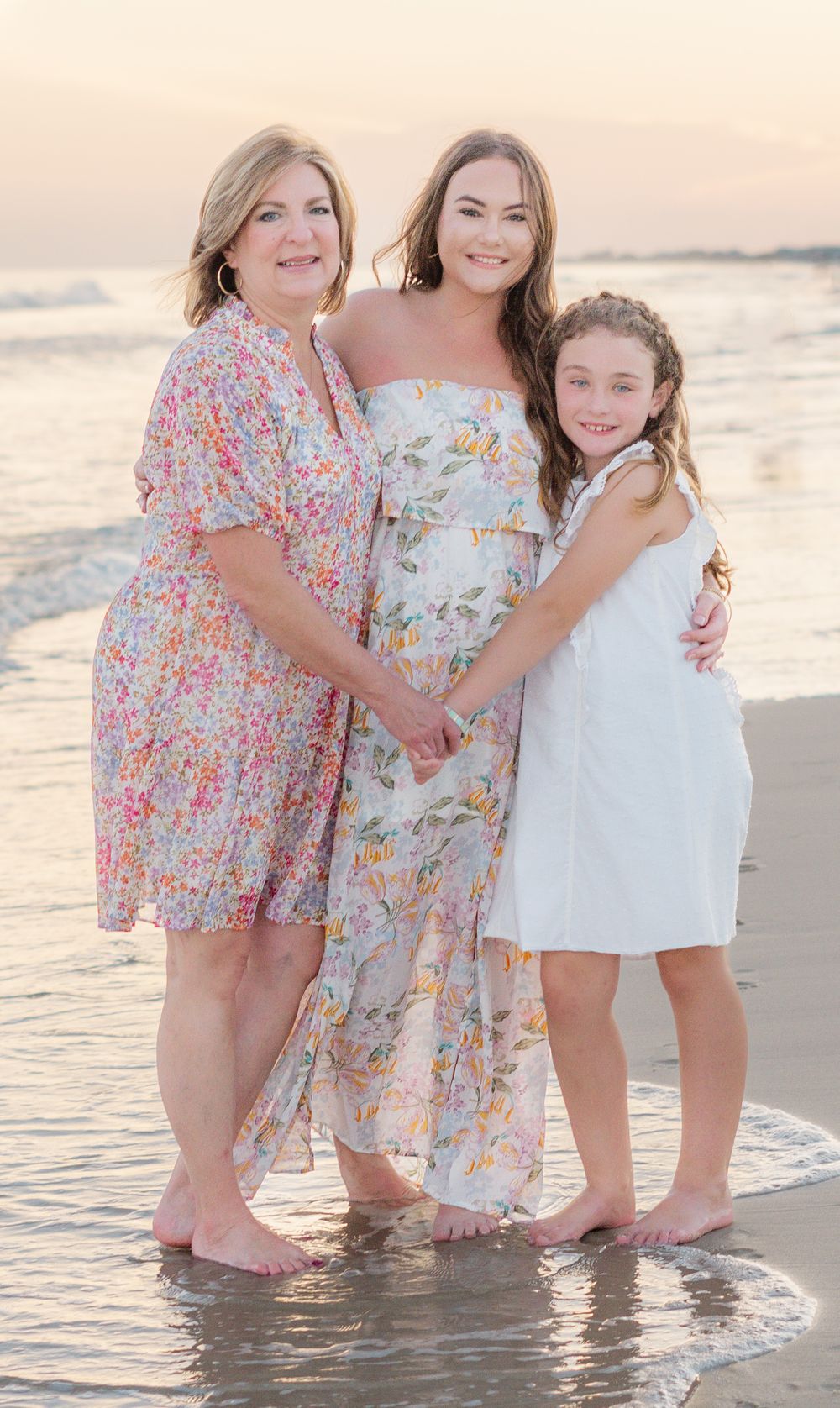 Three generations pose together on beach at sunset wearing floral and white dresses.