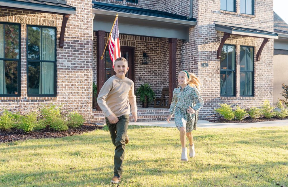Two figures running and playing on a lawn in front of a brick house with an American flag.