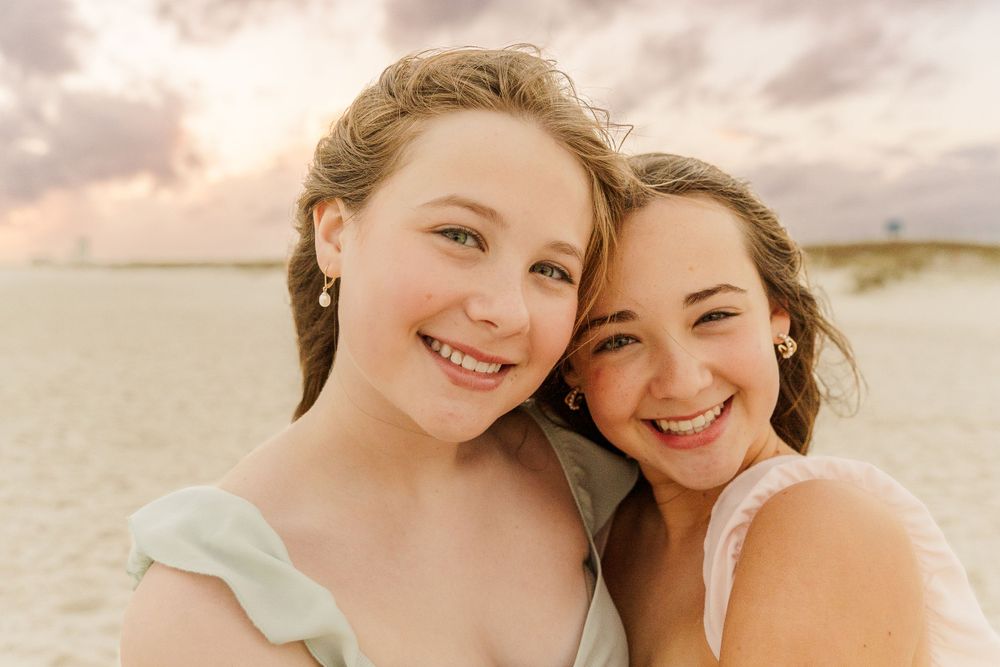 Two friends smiling together at sunset by the beach in pastel dresses.