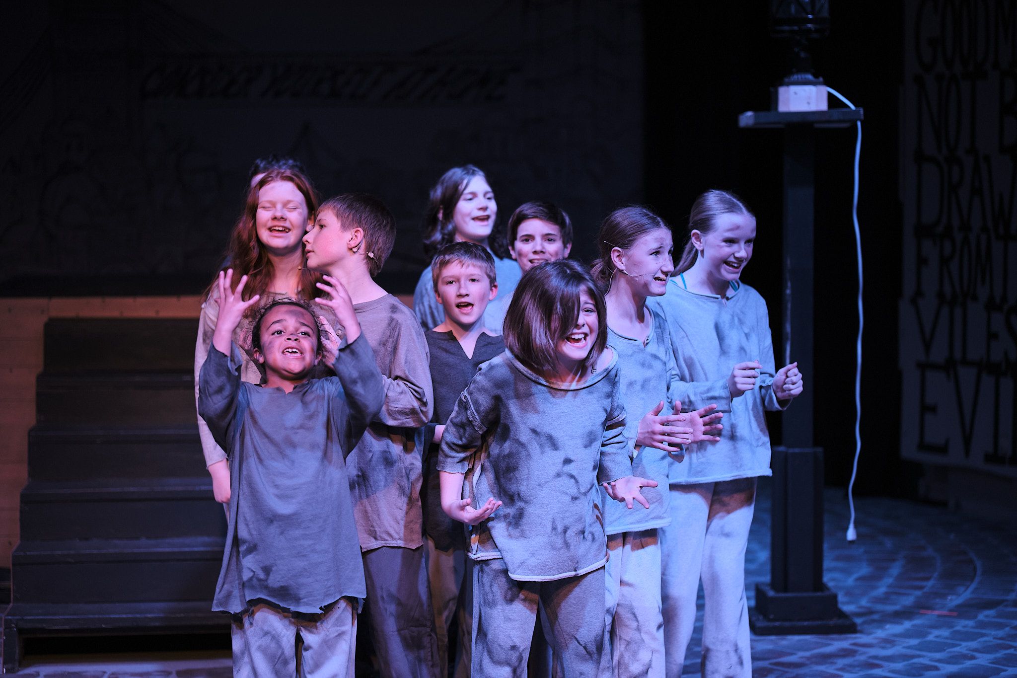 Theater performers in gray costumes gather under stage lighting during dramatic performance.