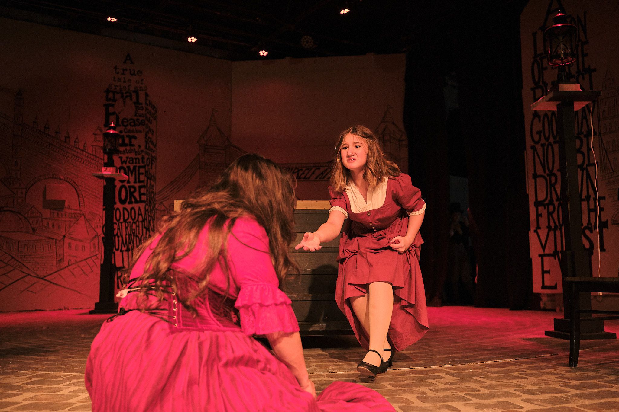 Two performers in red period dresses act out dramatic scene on theatrical stage.