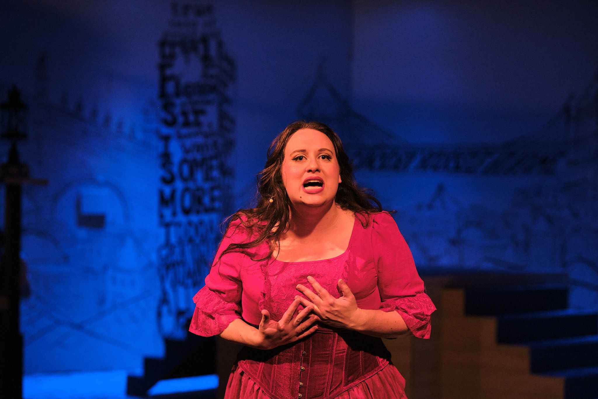 Performer in red dress sings passionately during theatrical production with blue lighting.