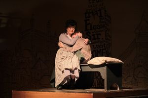 Actor in historical costume performs emotional scene on dimly lit stage with rustic wooden furniture.