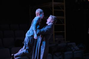 Two performers in period costumes engage in dramatic scene under blue lighting on dark stage.