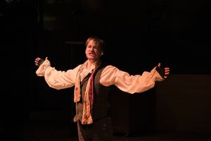 Stage performer in flowing white shirt gestures expressively during theatrical performance.