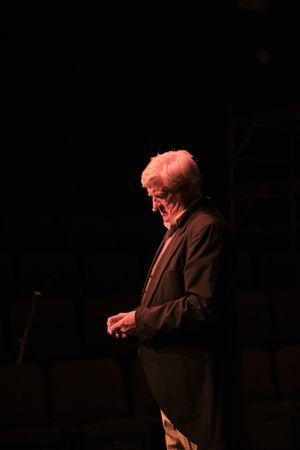 Side-lit performer in dark suit stands thoughtfully on darkened stage.