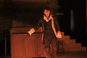 Performer in period costume stands by wooden furniture on dimly lit stage.
