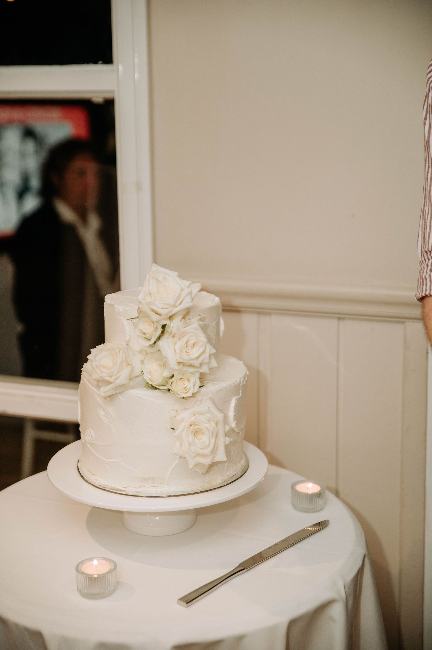 Elegant white wedding cake decorated with fresh roses sits on a table with candles.