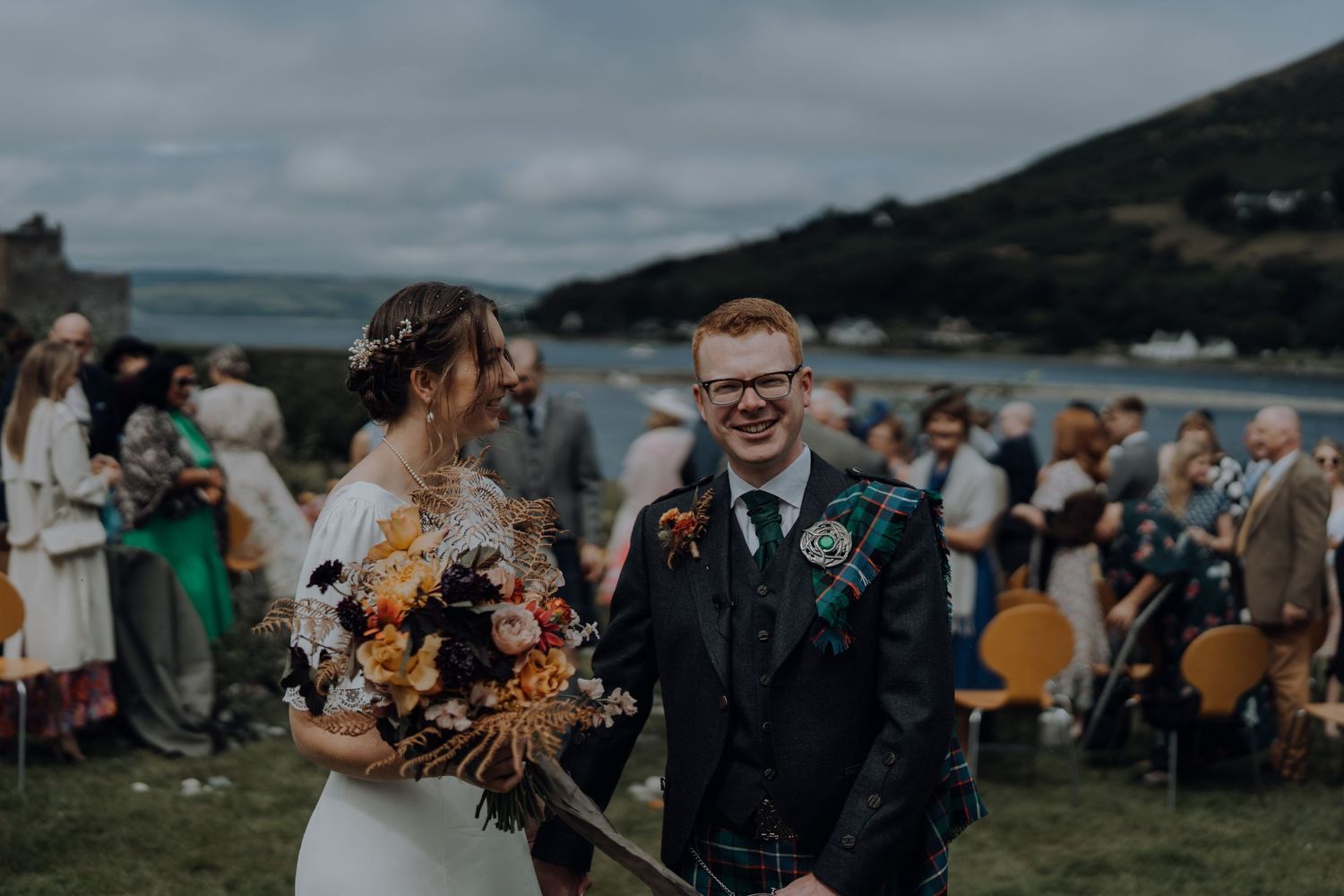 Coastal wedding ceremony with couple and guests gathered on hillside overlooking ocean.