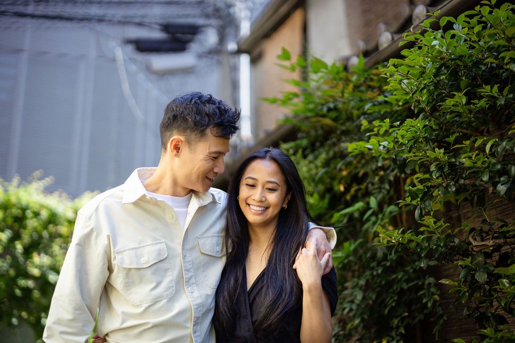 A couple share an intimate moment while standing next to green foliage in casual attire.