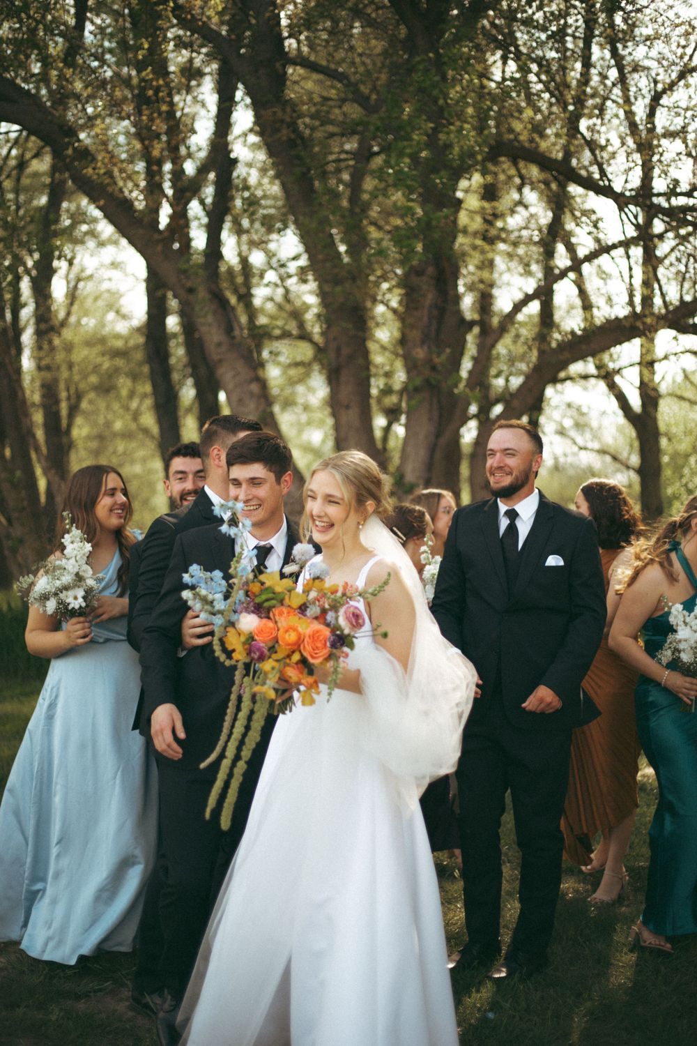 A wedding party poses together in a wooded setting with colorful bouquets and formal attire.