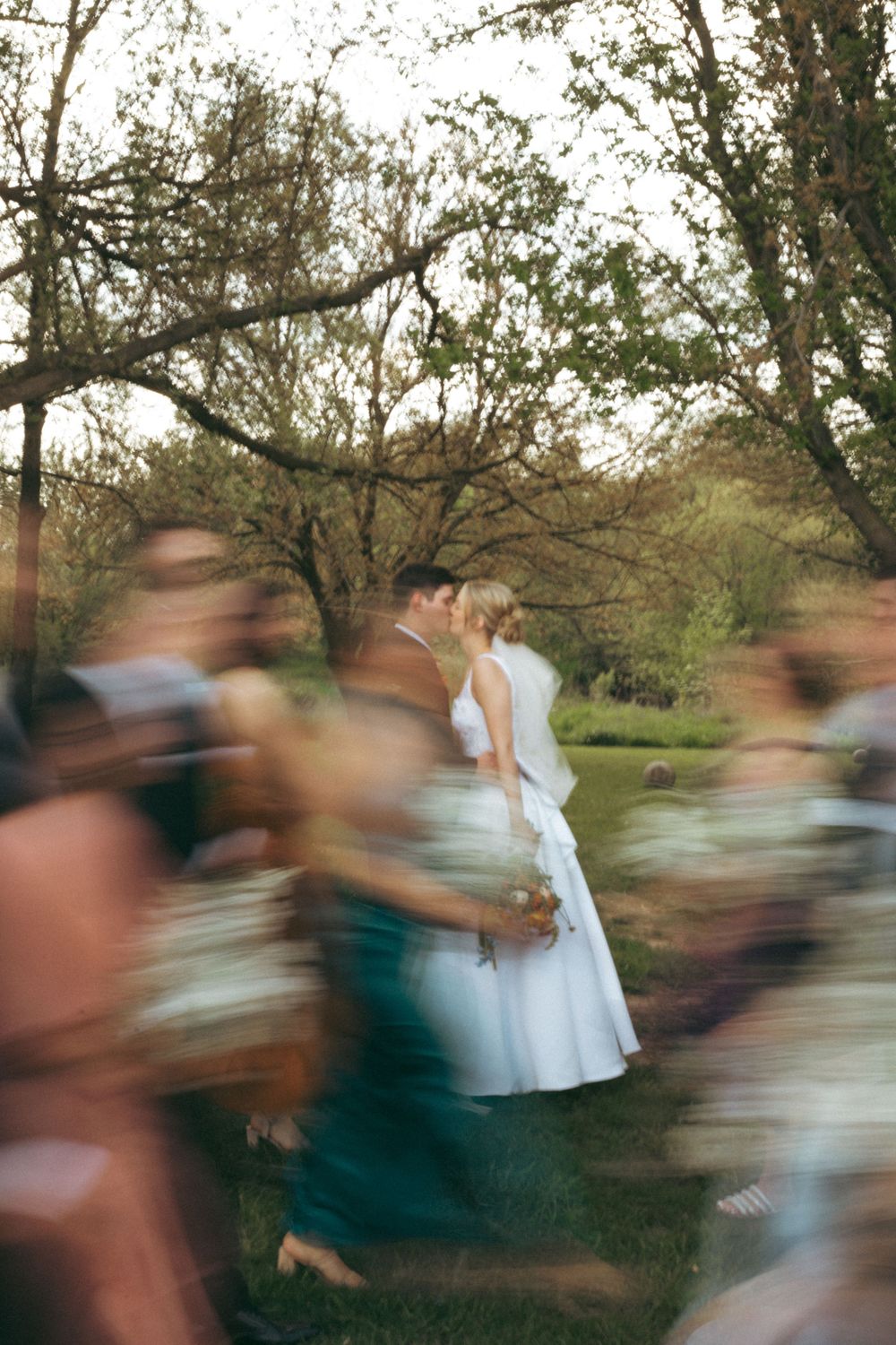 A wedding couple stands still while guests create motion blur around them in a forest setting.