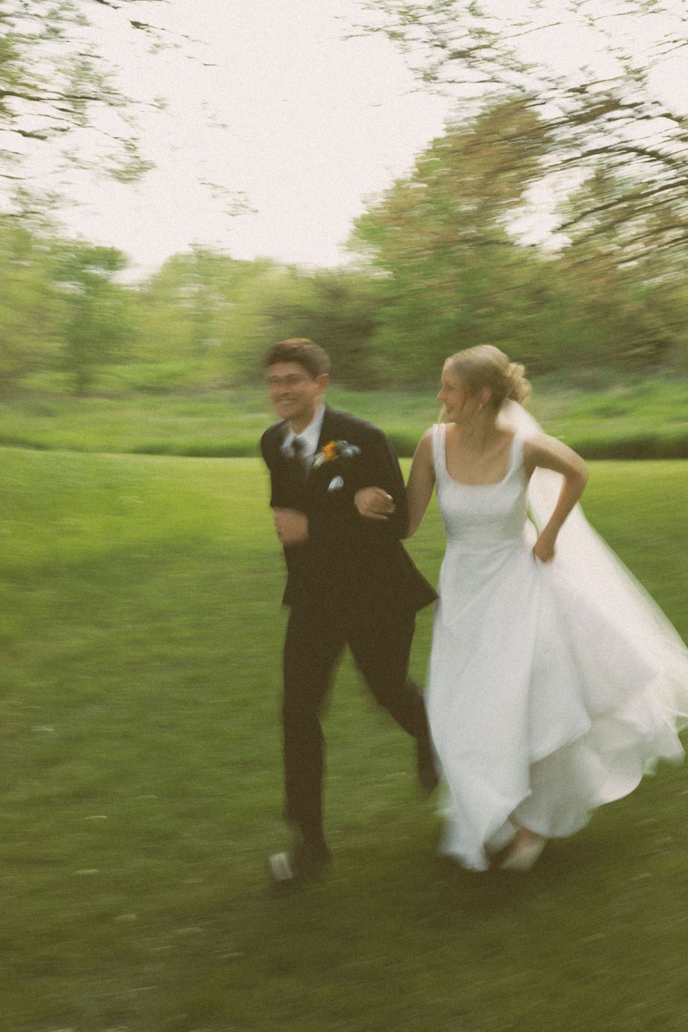 A couple runs playfully through a green field in wedding attire.