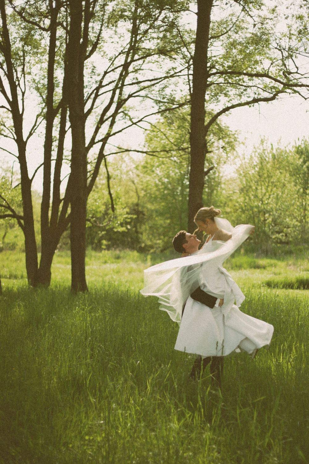 A joyful couple spins in an embrace amid tall grass and trees.