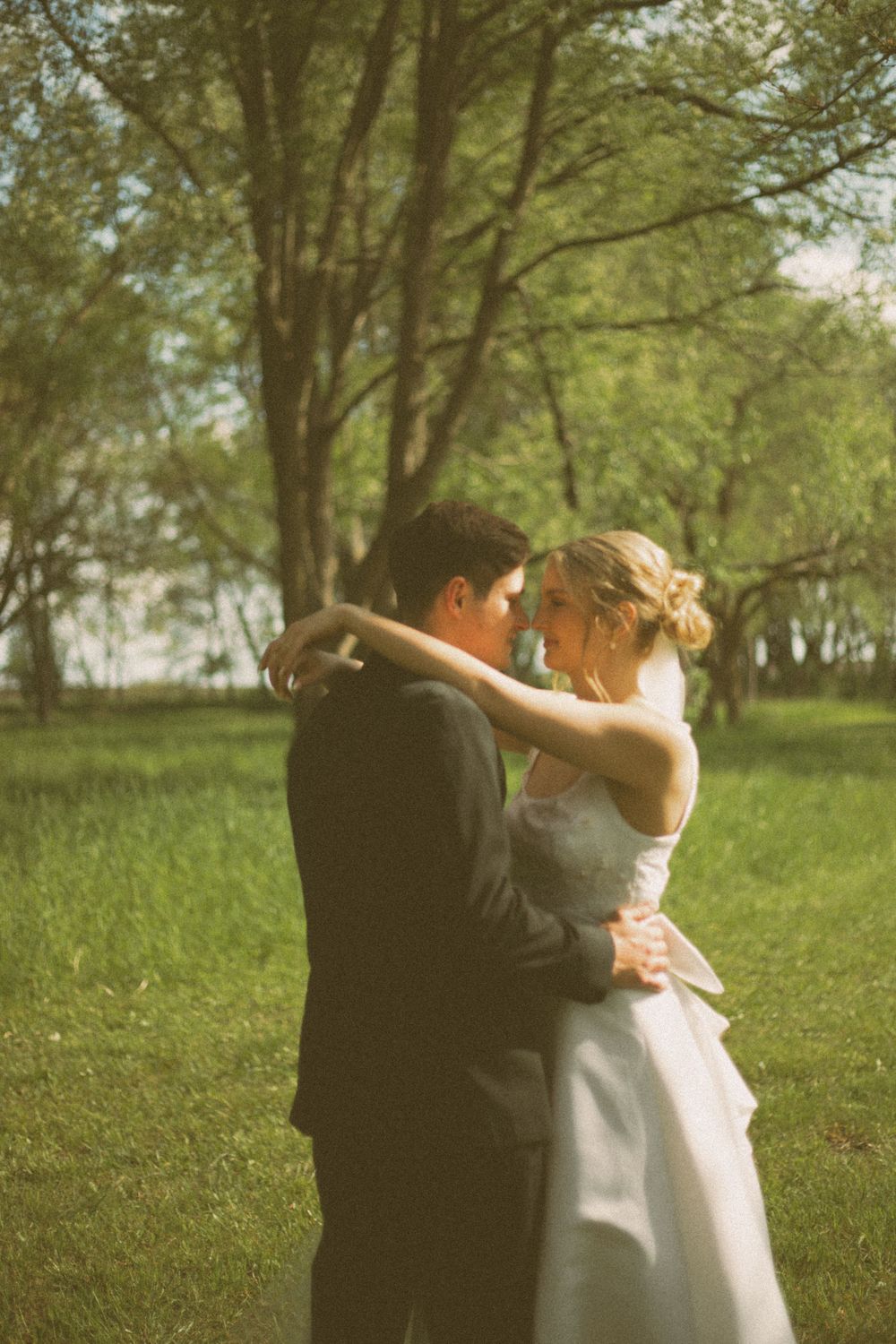 A romantic outdoor wedding portrait showing a couple embracing on lush green grounds with trees in the background.