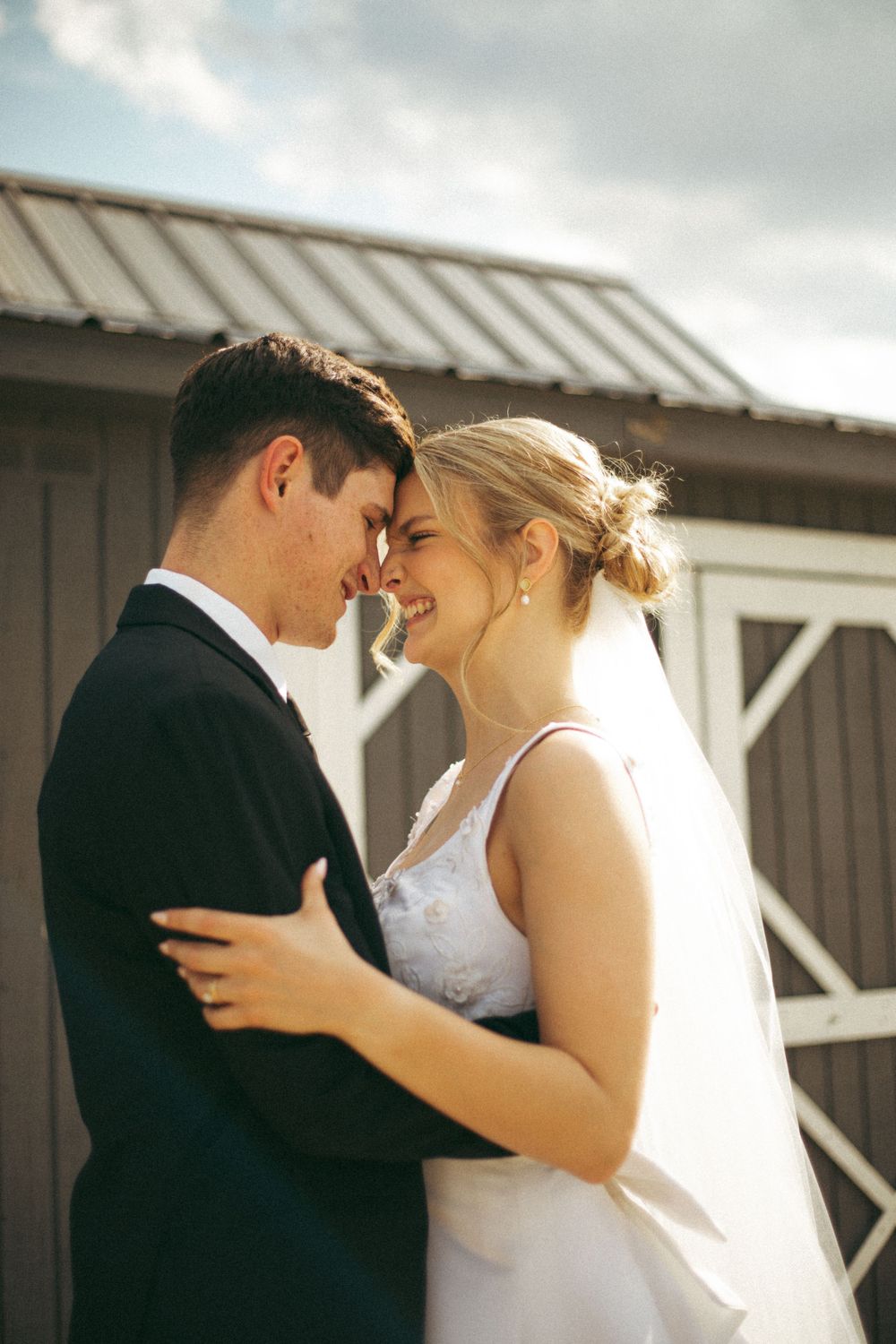 A sun-lit wedding portrait showing a tender moment between newlyweds outside a barn.