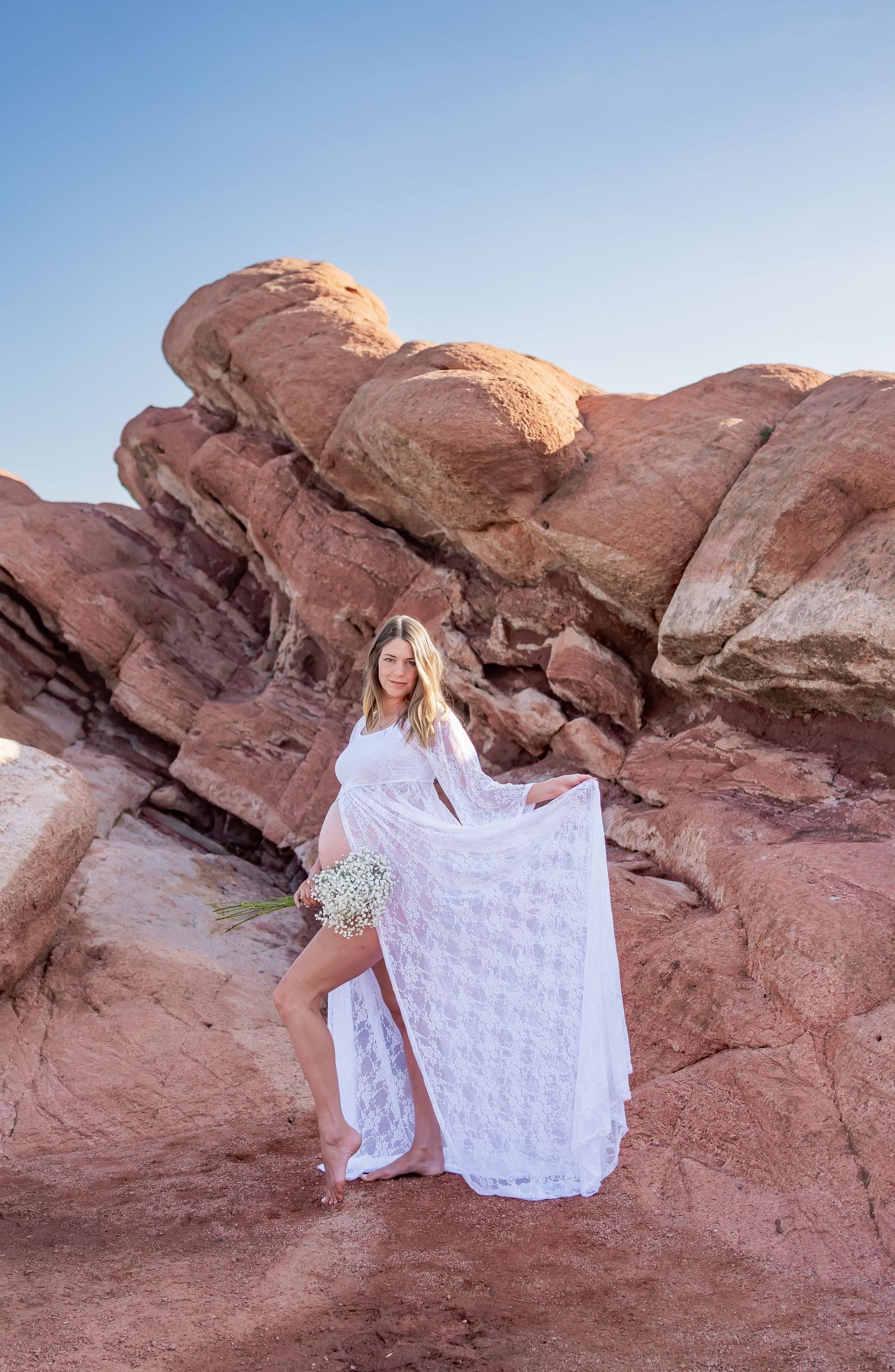 An elegant maternity portrait against red rocks with flowing white lace dress and baby's breath flowers.