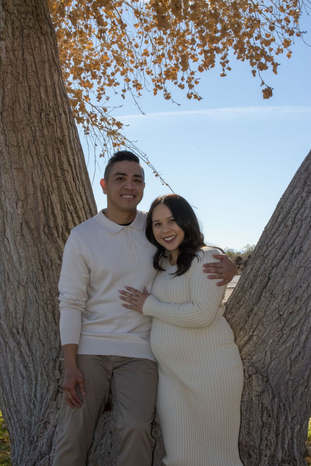 Smiling couple in matching cream outfits posing between two large trees with golden fall leaves.