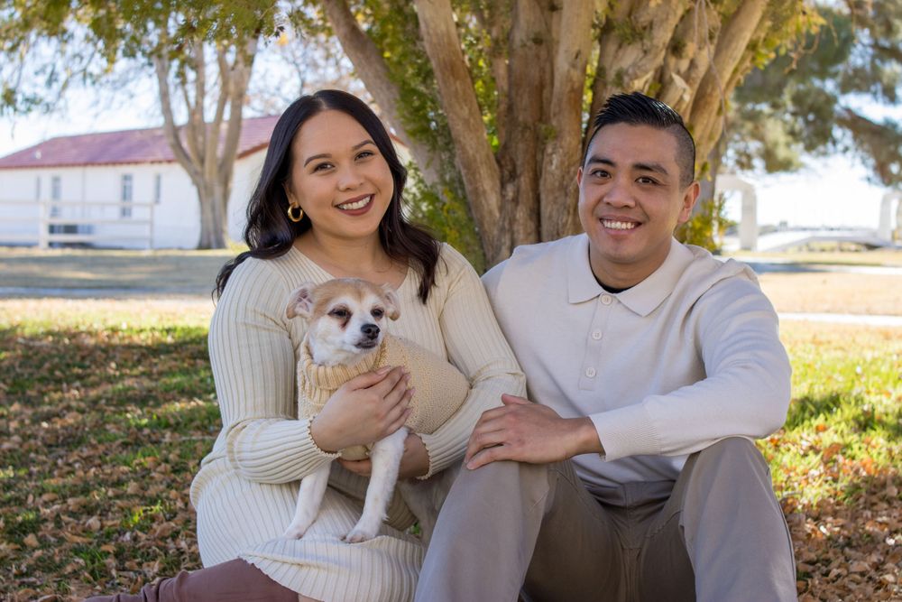 Happy couple in cream sweaters sitting on grass with their small Chihuahua dog under a tree.