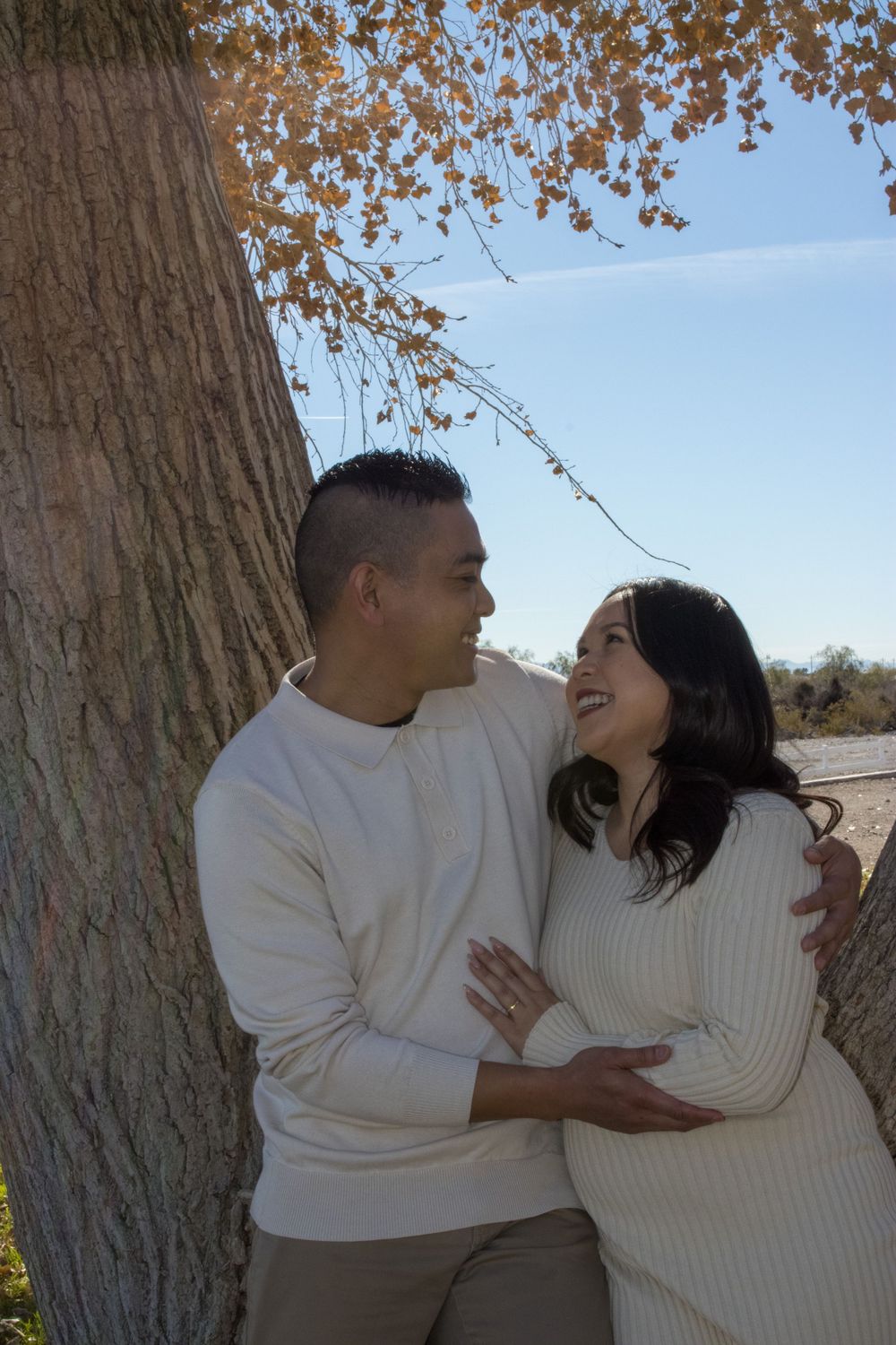 Couple in cream sweaters sharing a romantic moment leaning against a large tree with autumn foliage.